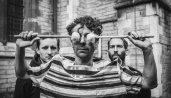 The trio pictured with drummer holding soft head sticks over his eyes