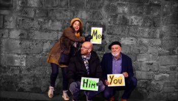 Three band members holding placards saying 'Me', 'Him', 'You'.